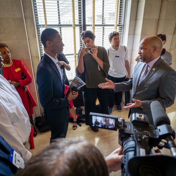 A student and legislator talk in the hallway surrounded by people and a camera with a microphone.