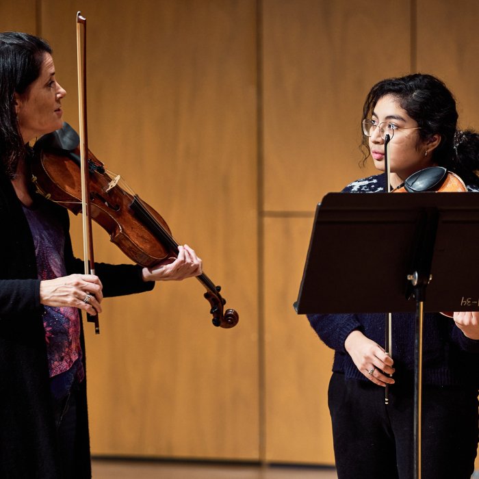 Female student playing violin listens to female instructor.