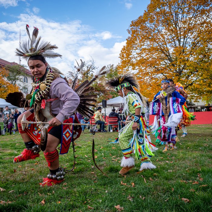 Native American men and women in traditional costumes dance outdoors while spectators watch.