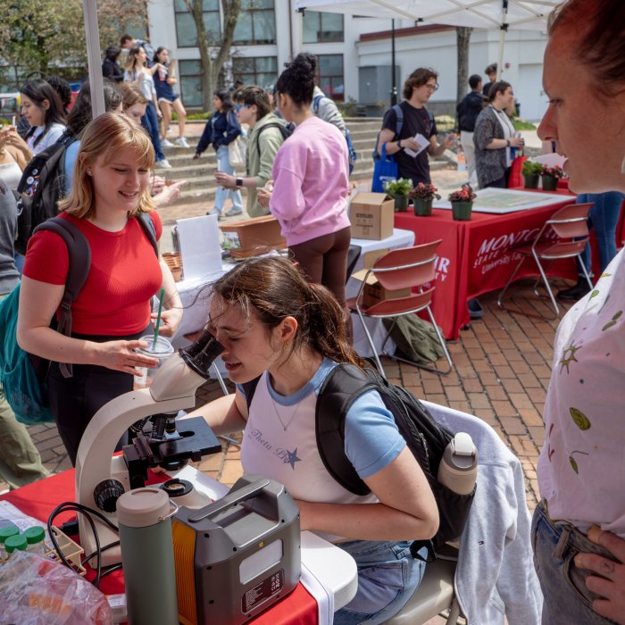 Two female presenting students participating in an Earth Day activity. One is seated looking through a microscope while the other stands and looks on.