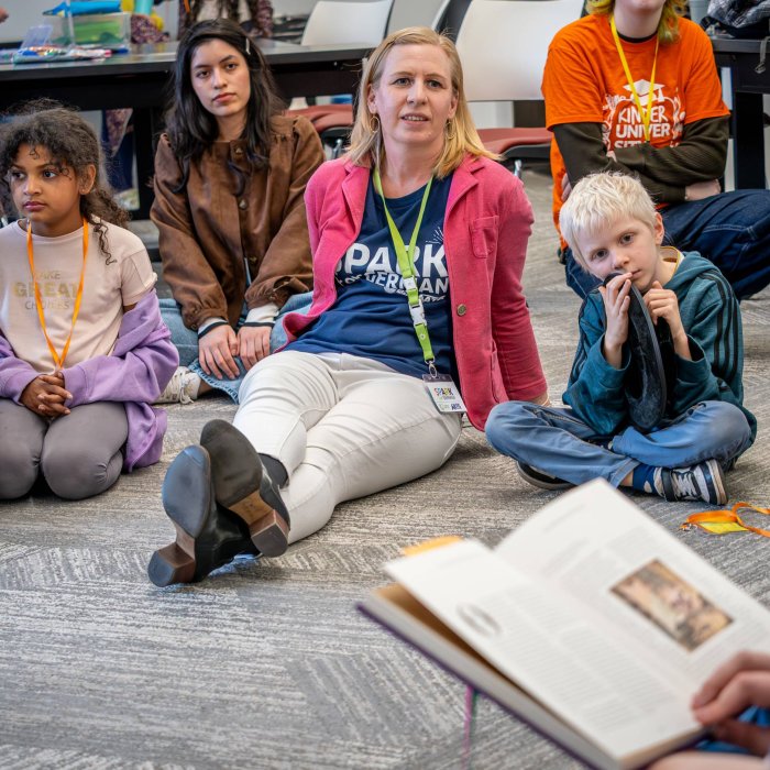 An adult sits with a group of childeren listening to someone reading a book