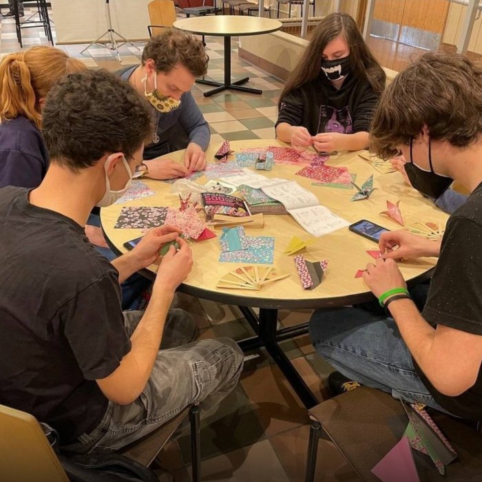 A group of six college students wear face masks and sit aorund a round table and fold oragami