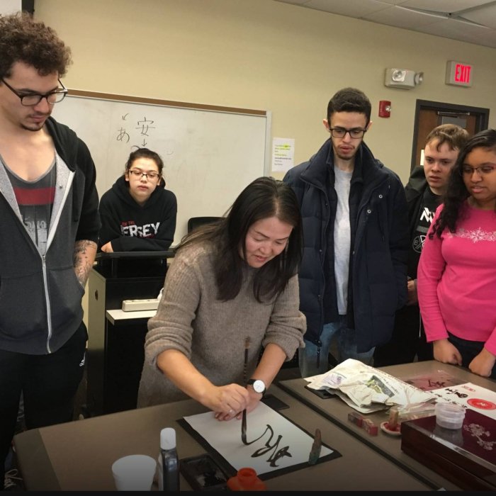 A woman demonstrates caligraphy on a sheet of paper while a group of college students watch