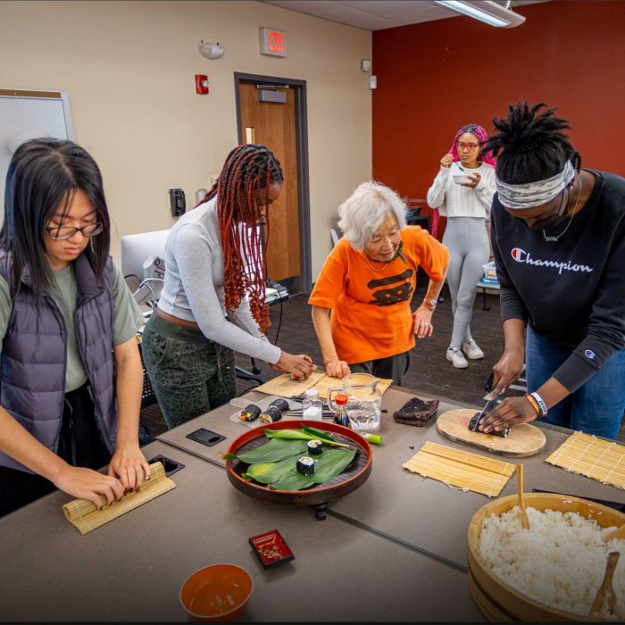 A group of three college students stand aorund a table and roll sushi as an older woman oversees their work