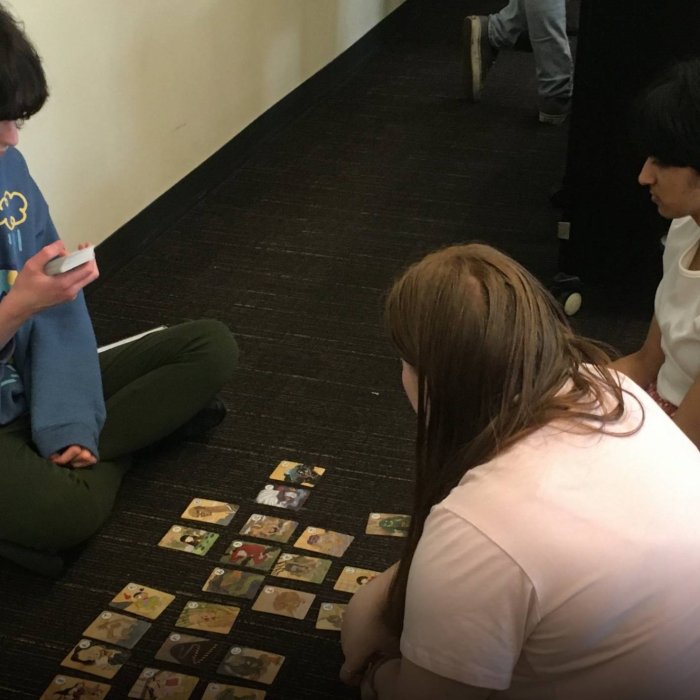 Two college students sit across from one student holding a deck of cards with a four rows of cards placed between them
