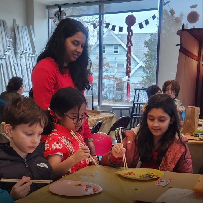 A woman watches as three children attempt to pick up M&M's with chopsticks