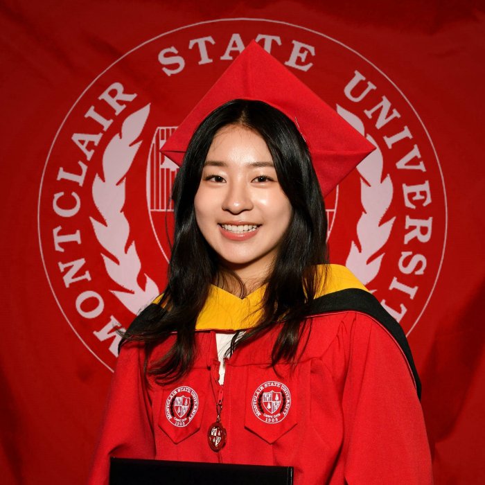 A picture of a student in their Montclair regalia in front of the Montclair logo. The student is holding their diploma.