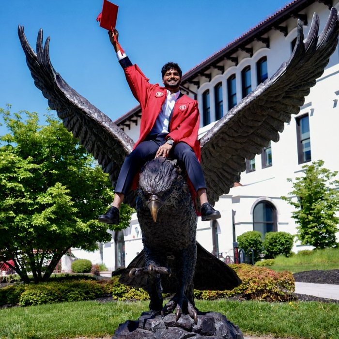 A picture of a student in their Montclair graduation gown sitting on top of the Red Hawk statue. The student is holding up their graduation cap in the air.