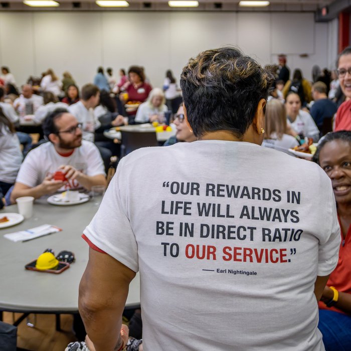 A person with a quote on the back of a T-shirt speaks with a colleague.