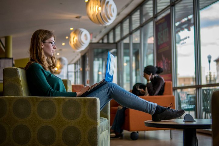 Student sitting in a lounge area, working on a laptop near large windows