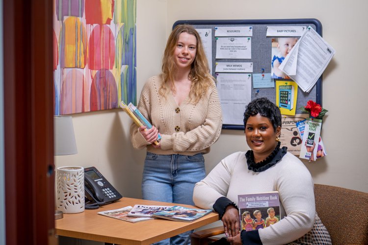 One woman stands and another sits while holding books.
