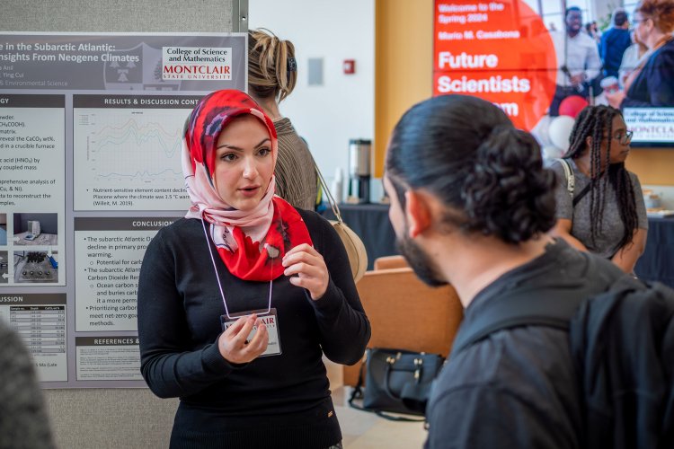 Busra Anil, a Montclair State University student wearing a red and white patterned hijab and black sweater, speaks with a guest during the Spring 2024 Mario M. Casabona Future Scientists Competition. She stands in front of her research poster on carbon sequestration, explaining her findings.