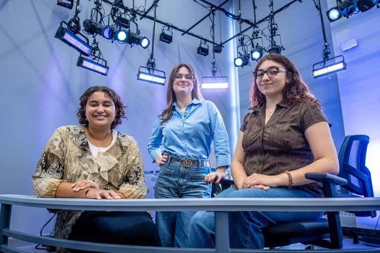 In a newsroom, three women, two seated at a desk and one standing between them pose with studio lights shining behind them.