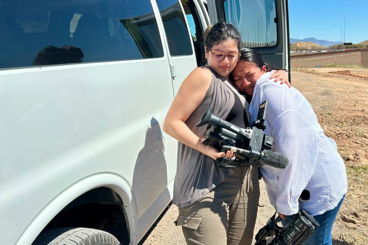A student holding a video camera hugs another student in the desert.