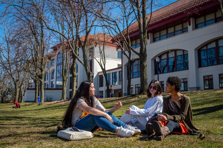 A group of students sitting outside on the grass on Montclair's campus. A campus building is in the background.