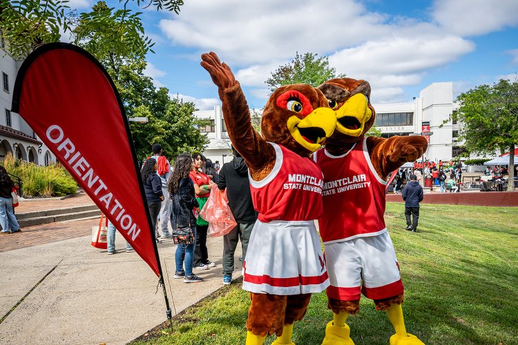 Mascots Roxy and Rocky standing outside with by the Orientation flag