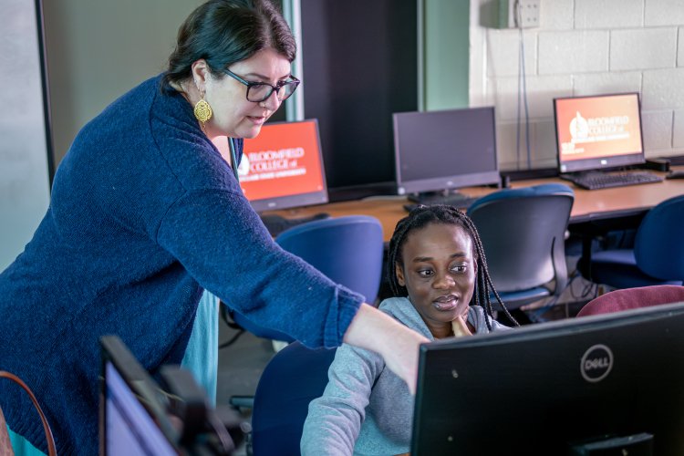 Woman pointing at a computer screen while woman sits in front of the screen