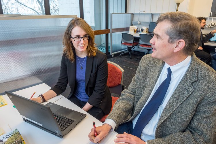Two people sit around a laptop at a desk in an office environment