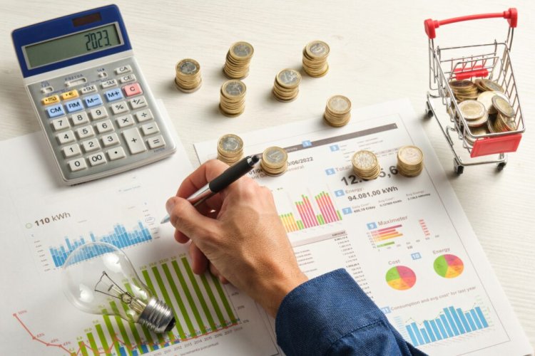 A hand holding a pen on a desk with assorted items related to billing (calculator, money, spreadsheet, etc.)