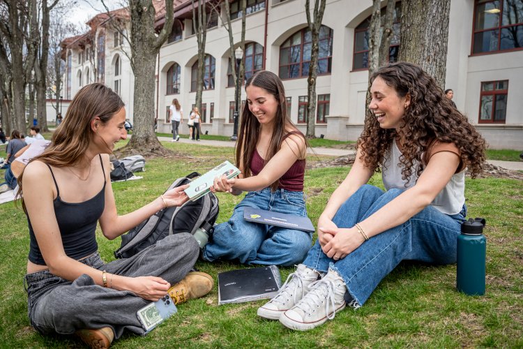 three students passing a notebook on the quad