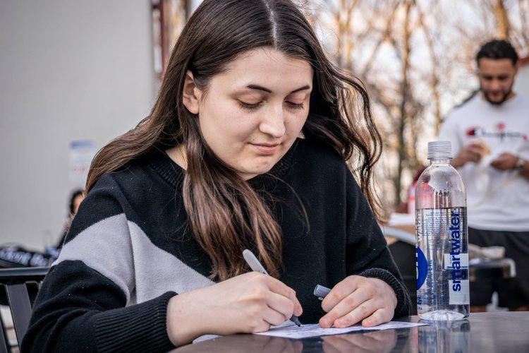 A college student sits at an outdoor table and writes on a piece of paper