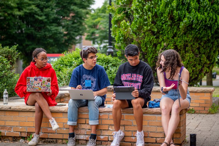 Four students sitting outside on a ledge with their laptops open chatting about school.