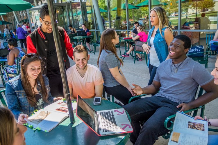 A group of college students sit around an outdoor table under an umbrella with laptops and books out