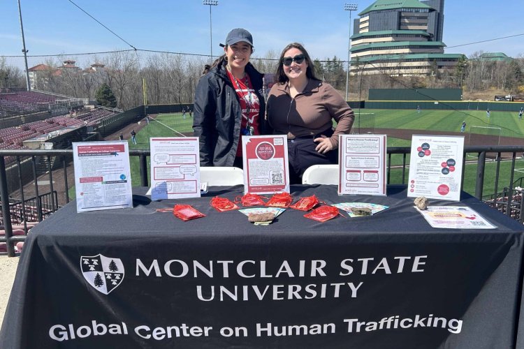 Two people stand at a baseball stadium, behind a table with tablecloth reading "Montclair State University Global Center on Human Trafficking"