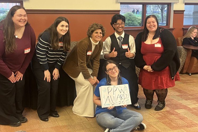 A group of six college students pose in a line, one students sits and holds a book which has the words "PPW Awards 2026" written in it 