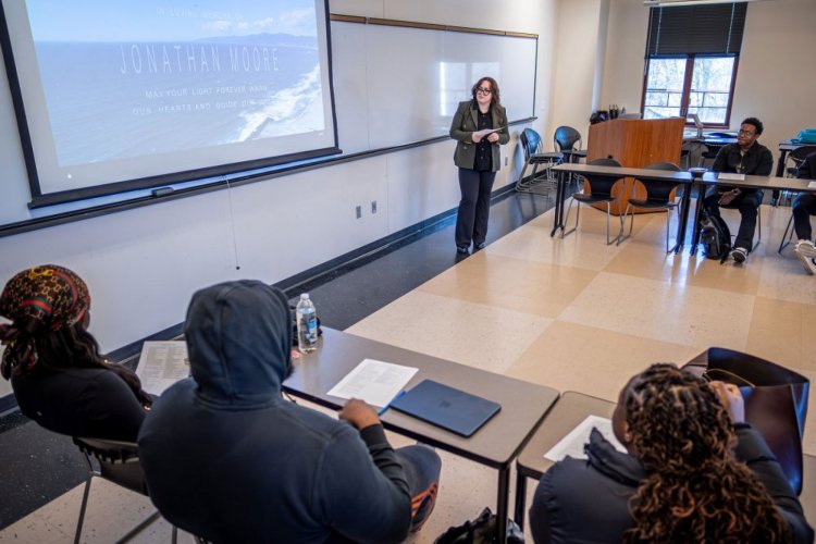A college student stands in front of a projector screen and presents to a class full of students