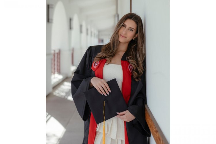 A college student in a graduation robe holds a mortarboard and leans against the wall of a courtyard