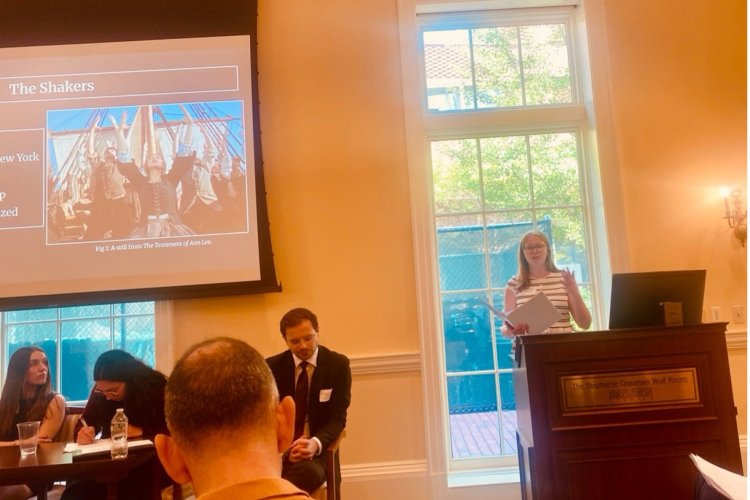 A woman stands at a dais and presents a PowerPoint to a room full of people