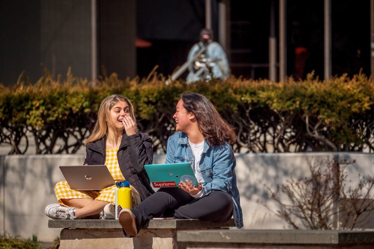 Two students sitting outside laughing with computers 