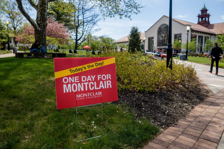 A red sign with while lettering says: One Day for Montclair. The campus is in background.