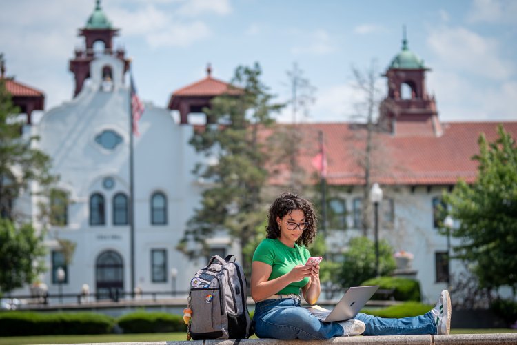 Woman sitting a ledge with laptop, phone, and bookbag. 