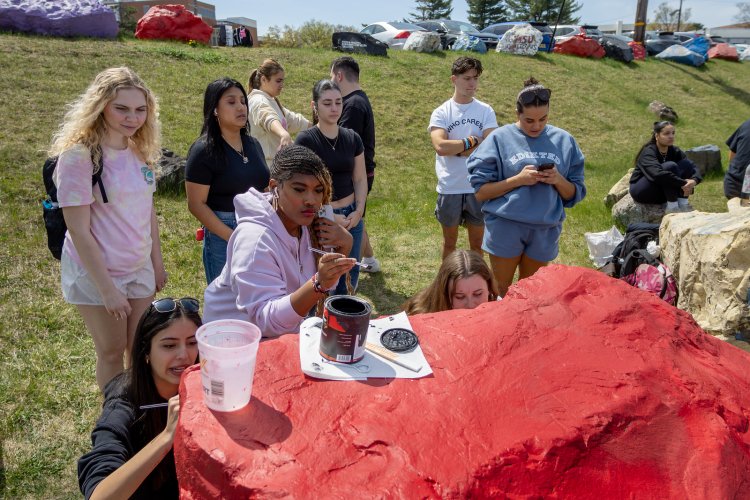 A group of students gathers around a large red campus rock, working together to paint it during an outdoor campus community event.