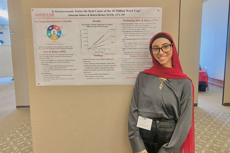 A college student wearing a black head covering and a long red scarf stands in front of a printed presentation pinned to a wall