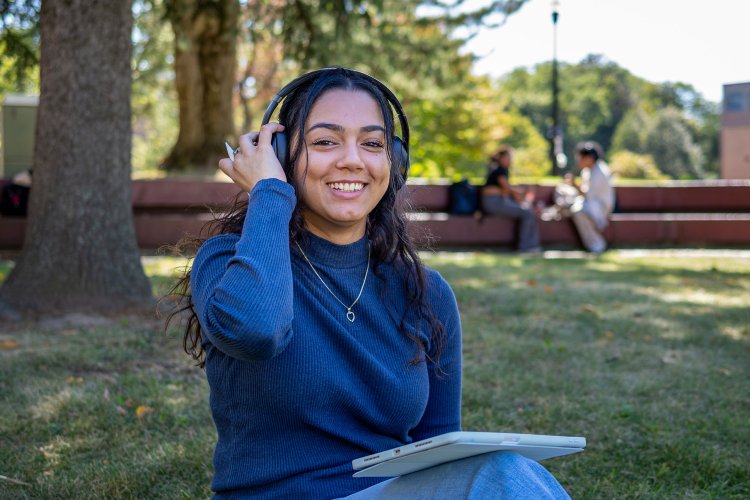 Student wearing headphones and smiling at the camera. They have an ipad on their lap.