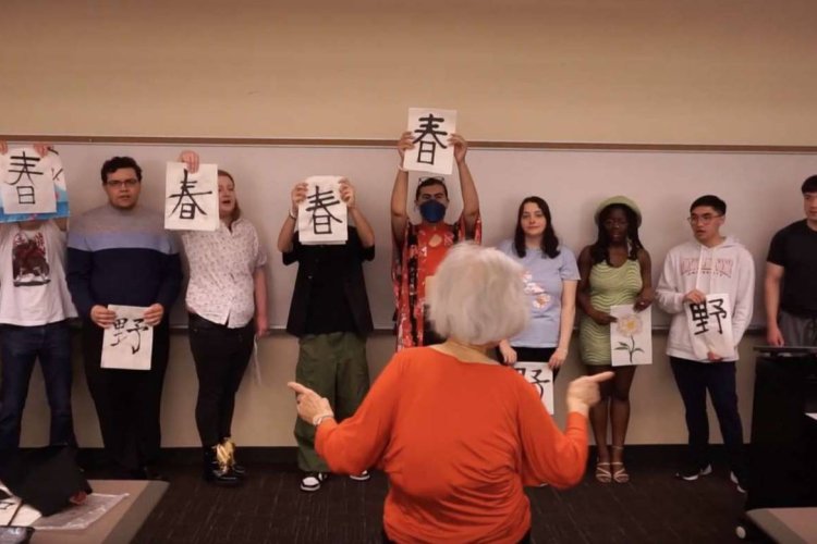 A group of college students stand in a line holding papers with Japanese vocabulary as they sing and follow an instructor's lead