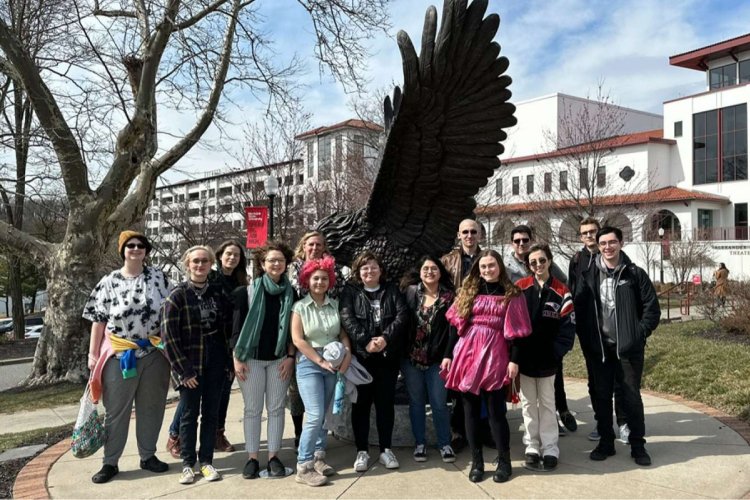 A group of college students pose together in front of a metal statue of a Red Hawk