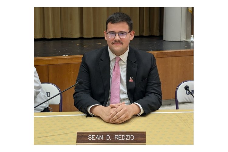A man wears a suit and tie and sits at a desk with a name plate in front of it