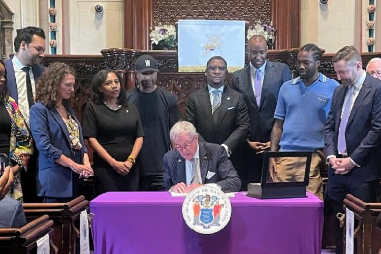 A group of people in professional clothing stand around a man seated at a table who is signing a paper