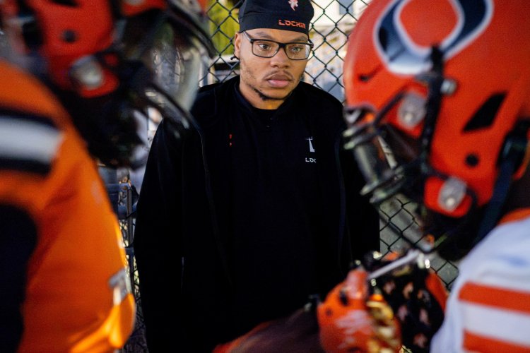 Montclair alumnus Emmanuel Ozias ’25, wearing a black Lock1n cap and jacket, stands by a chain-link fence surrounded by Orange High School football players in orange helmets and uniforms.
