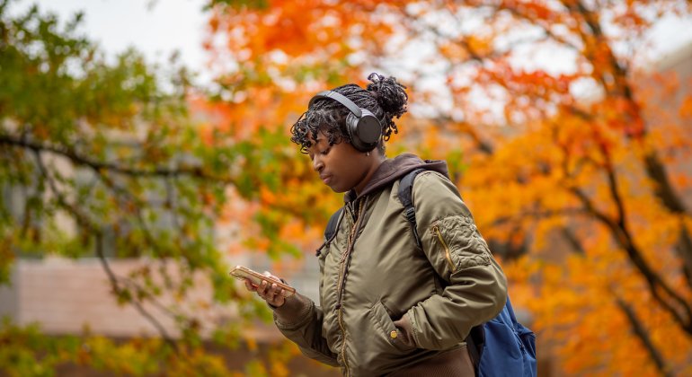 A student walking while looking at her phone with autumn trees behind them