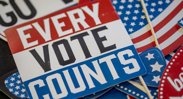 Close-up of colorful “Every Vote Counts” election signs and American flags promoting voter participation.