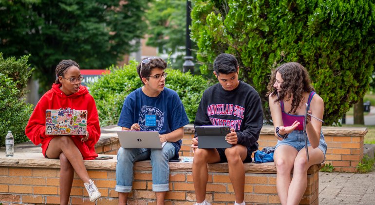 Four students sitting outside on a ledge with their laptops open chatting about school.