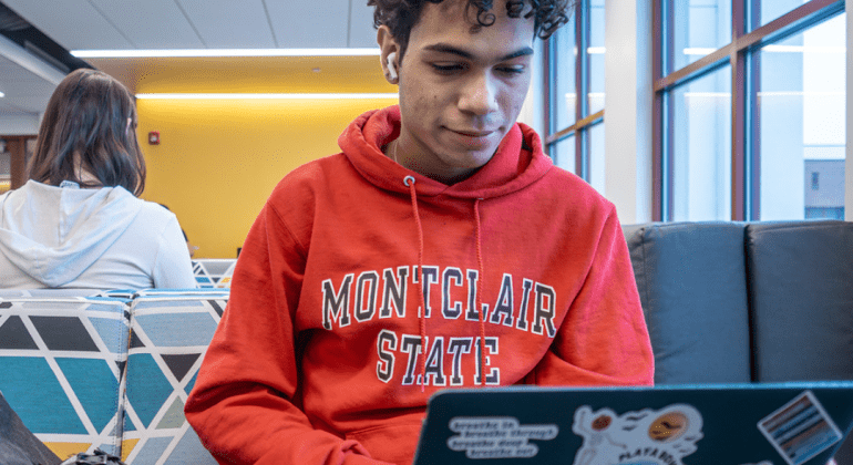 Student wearing red Montclair State hoodie studies on his laptop.