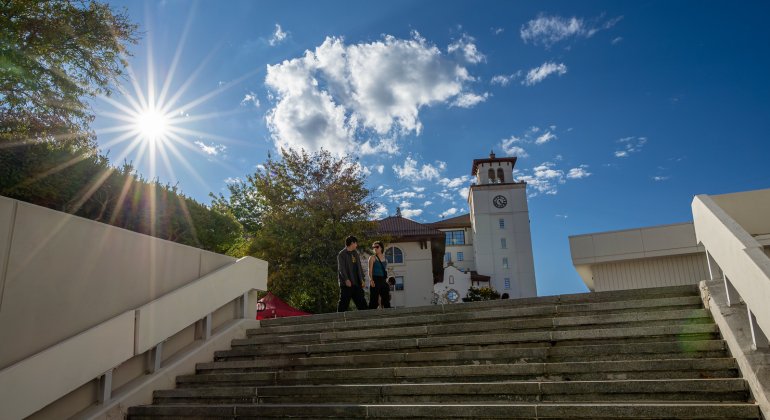 Two students walk up campus steps toward a clock tower under a bright blue sky with the sun shining through nearby trees.