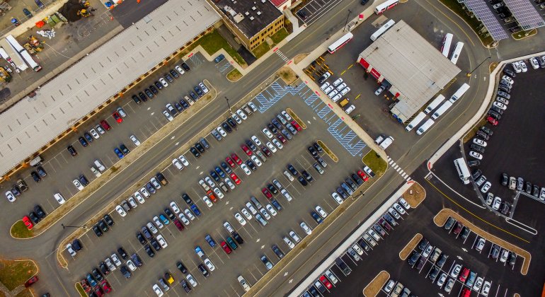 An aerial view of Montclair State University commuter parking lots shows parked cars, shuttle buses, campus roadways, and solar panel canopies.