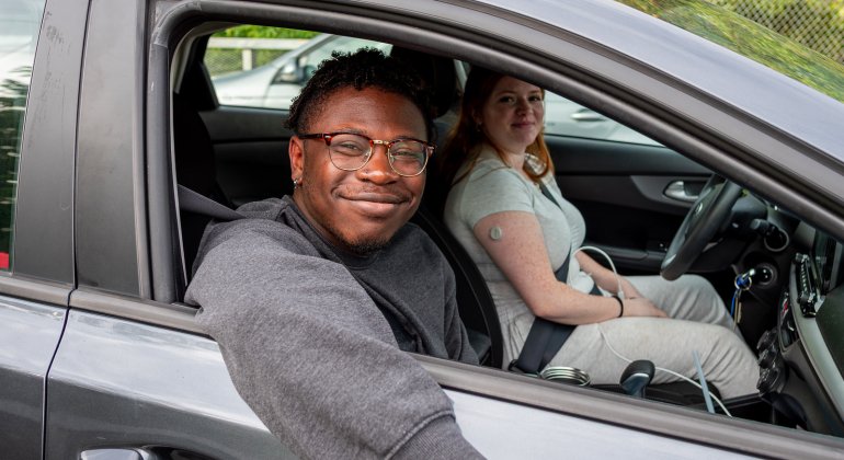 Two smiling college students sit in a parked car, with the front passenger leaning out the open window while the driver looks toward the camera.
